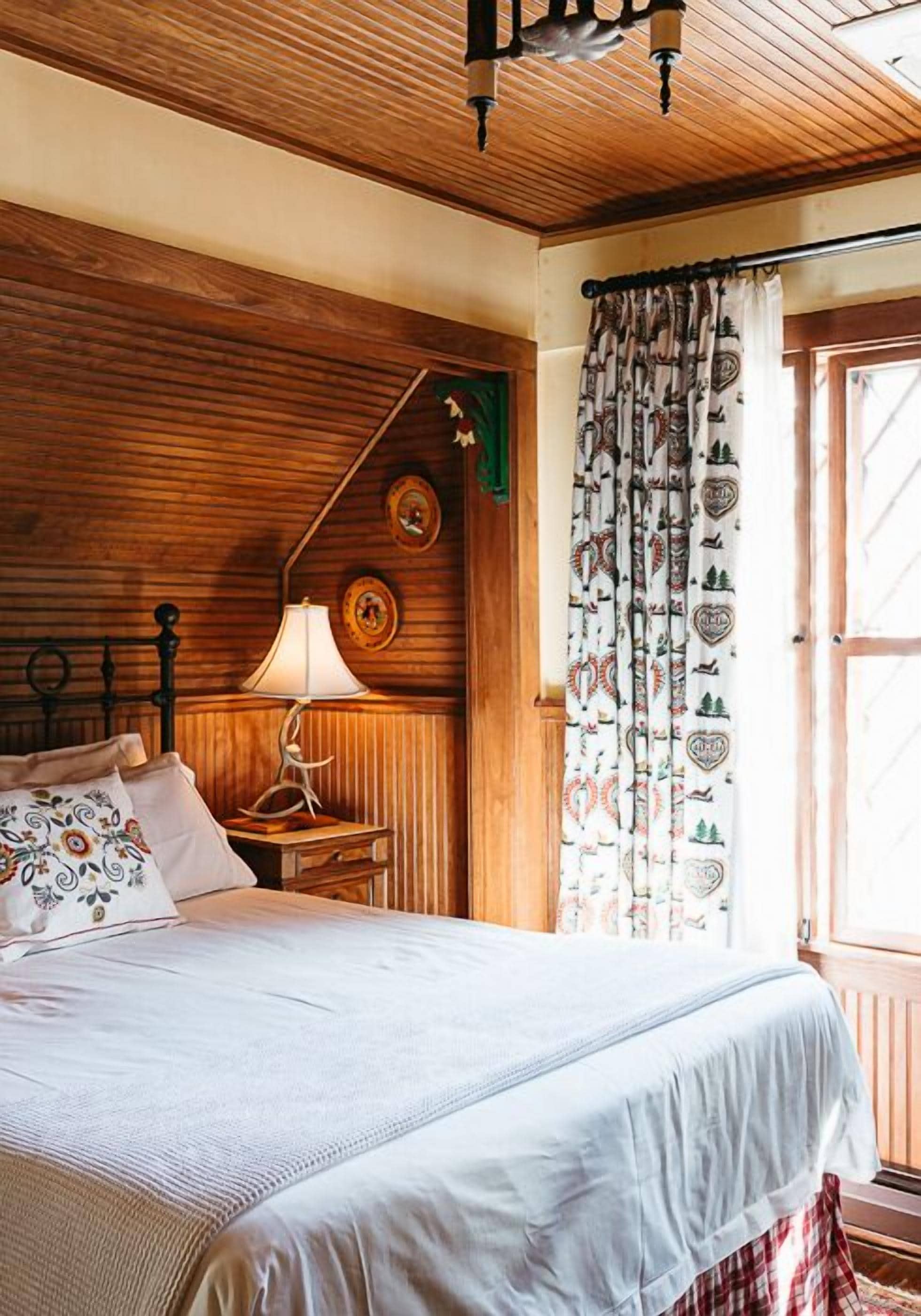 A wooden-paneled room with a bed, a nightstand, a lamp, and a window with patterned curtains.