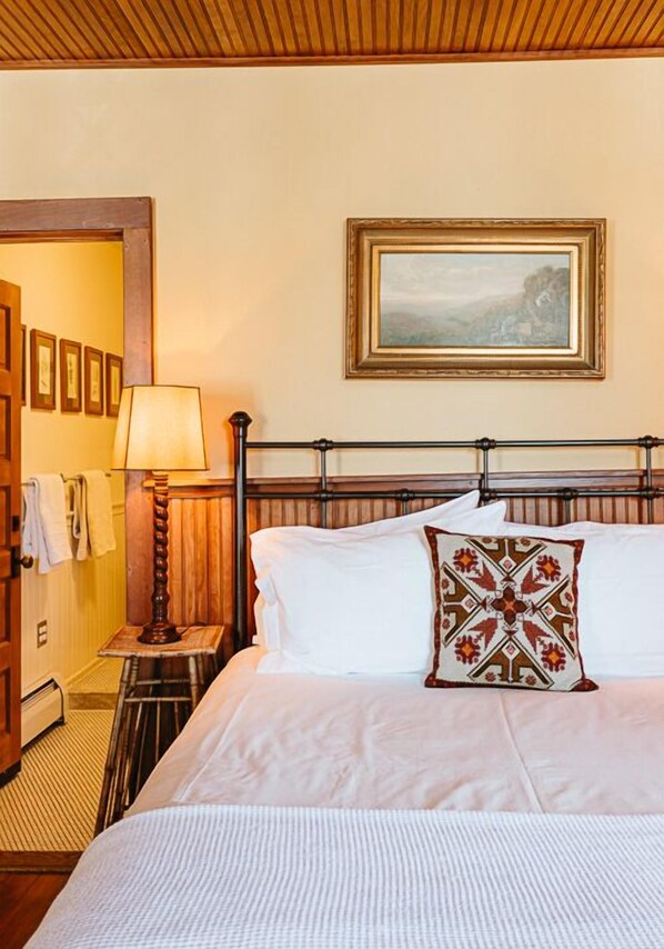A neatly made bed with white linens and a decorative pillow, a wooden headboard, a framed painting above the bed, and a lamp on a bedside table.