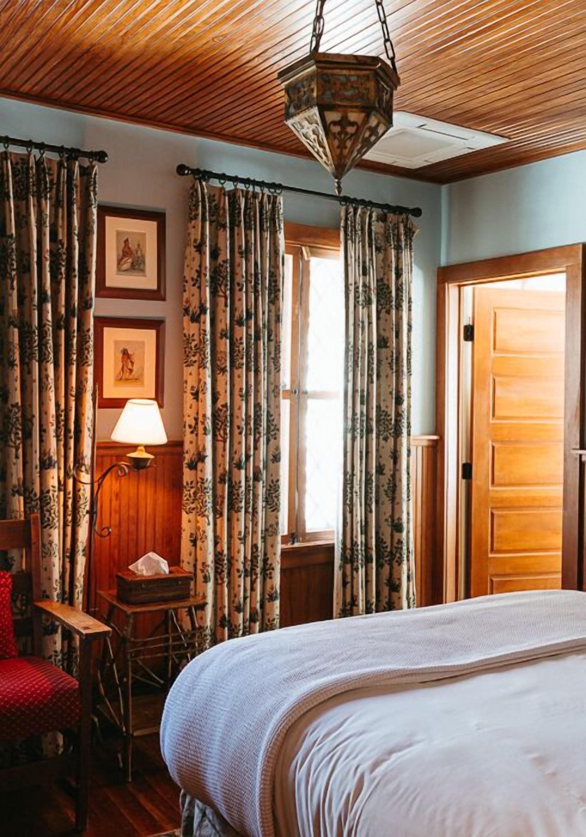 Una habitación con techo de madera, cortinas florales, una cama, un escritorio con lámpara y un cuadro en la pared.