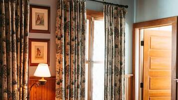 Una habitación con techo de madera, cortinas florales, una cama, un escritorio con lámpara y un cuadro en la pared.
