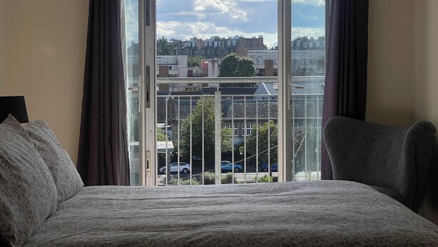 A bedroom with a large bed, a view of the city through an open window, and a balcony with a railing.