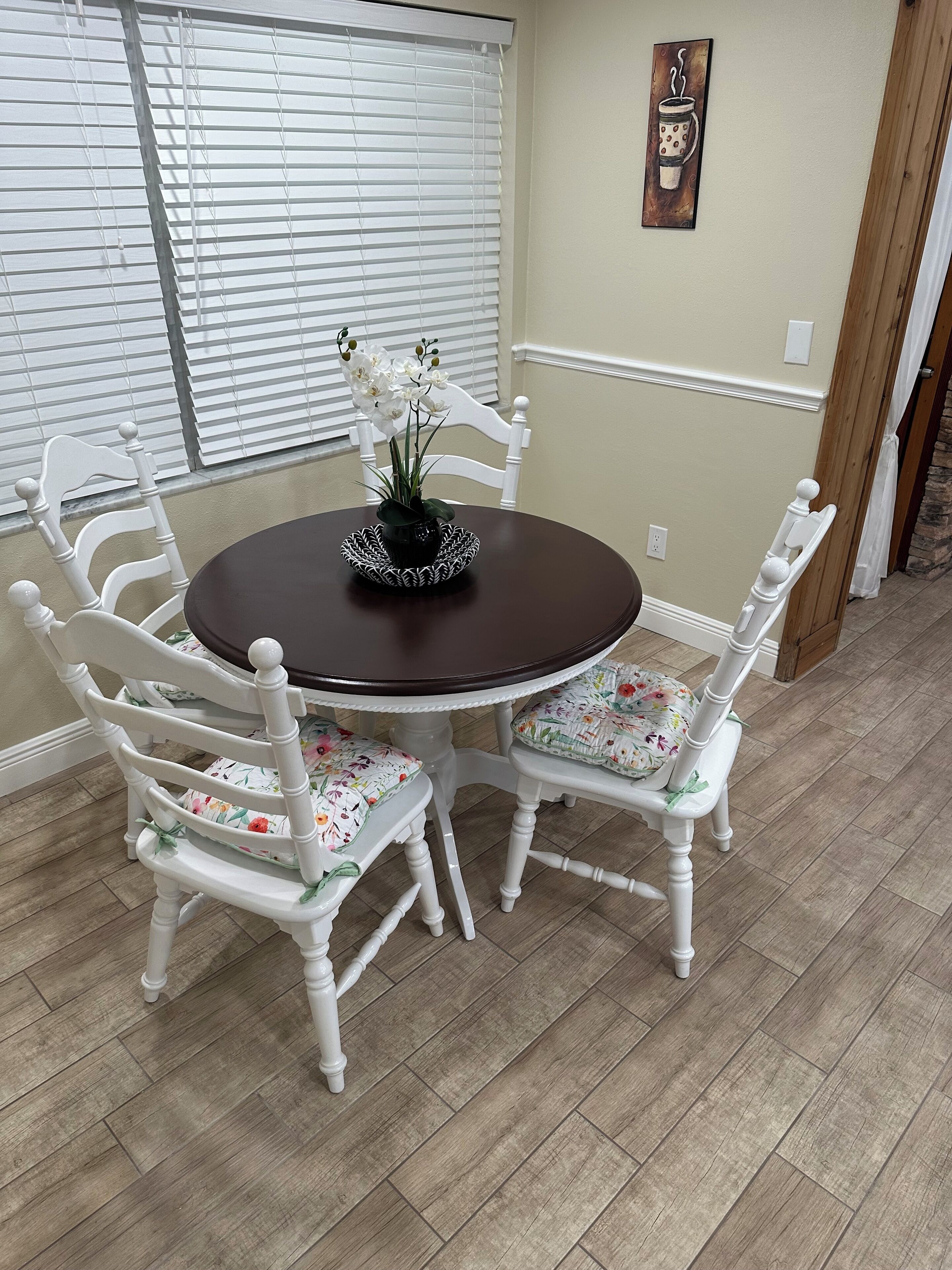 A dining area with a round table, white chairs with floral cushions, and a vase with white flowers.