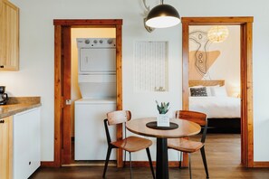 Una cucina compatta con forno a incasso, un'area pranzo con un tavolo rotondo e due sedie, e una camera da letto con un letto e un'opera d'arte appesa al muro.