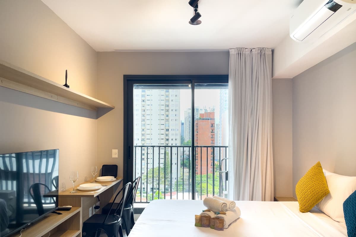 Hotelkamer met een bed, bureau met televisie, stoel, balkon met uitzicht op gebouwen en raam met gordijnen.
