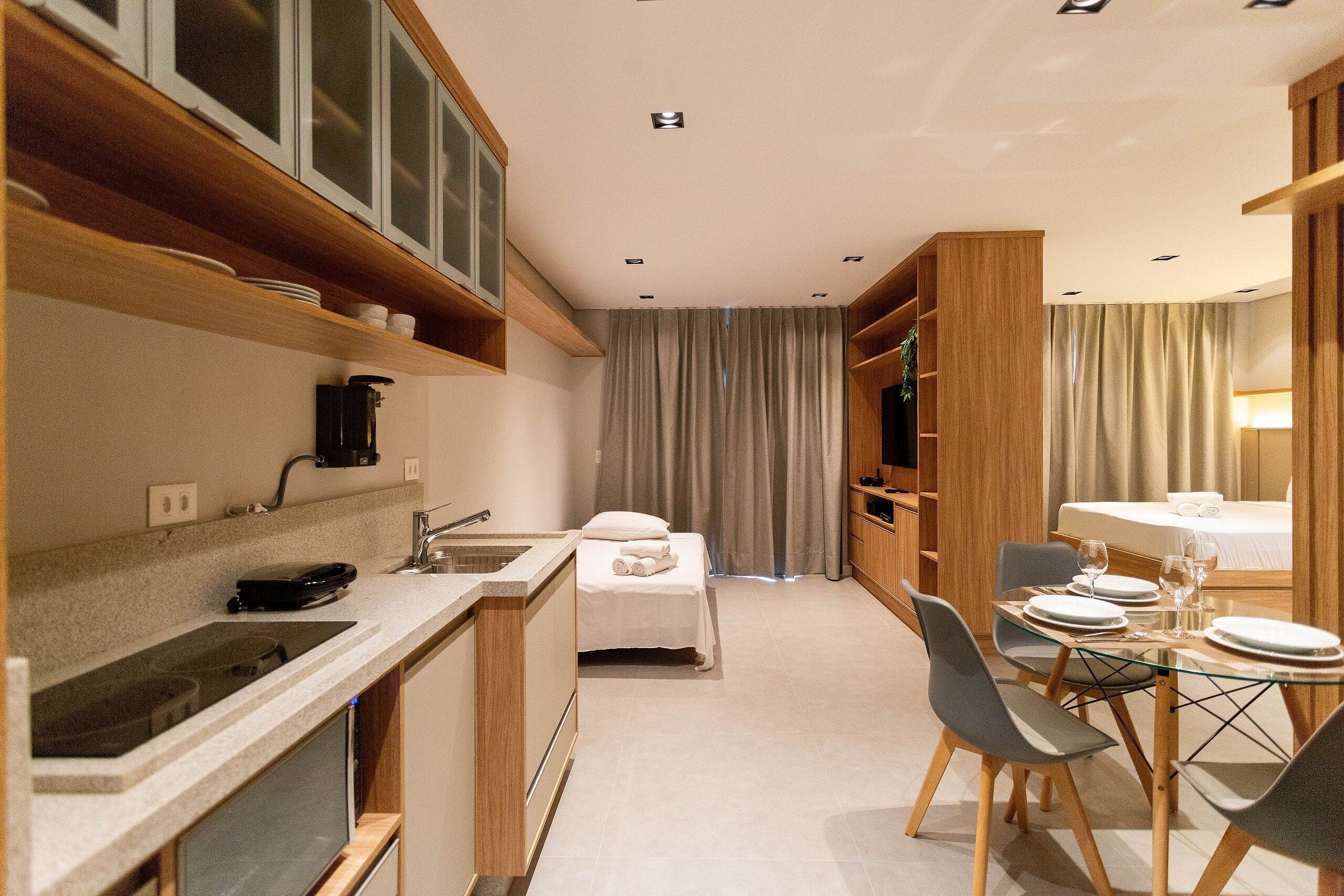 A compact hotel room with a kitchenette, a bed with a white duvet, a dining area with a glass table and chairs, and a large window with curtains.