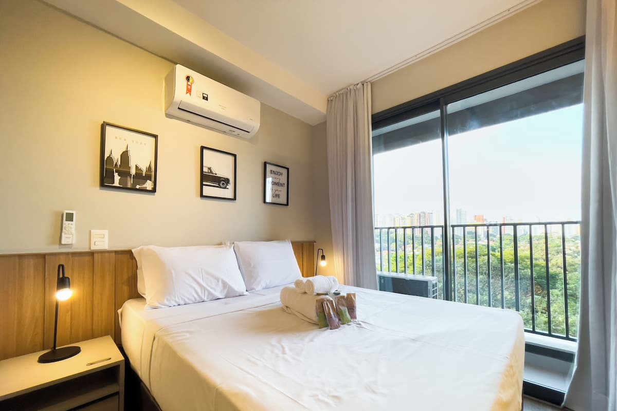 Hotelkamer met een bed, nachtkastjes, een airconditioner, ingelijste foto's aan de muur, en een balkon met uitzicht op gebouwen en groen.