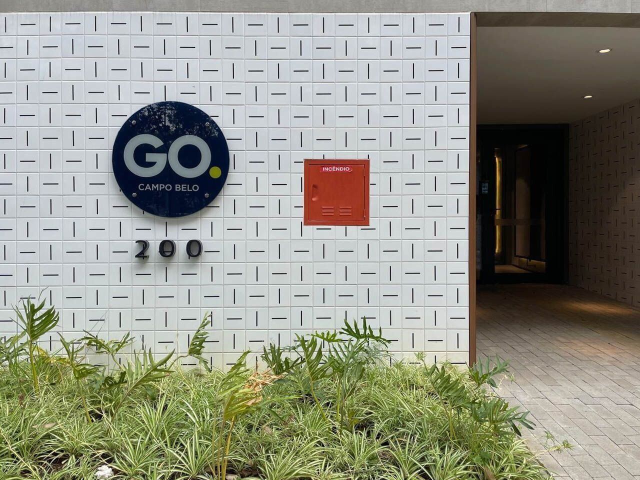 A modern building entrance with a white patterned wall, a blue circular sign with "GO CAMPO BELO", and a red sign labeled "INCÊNDIO".