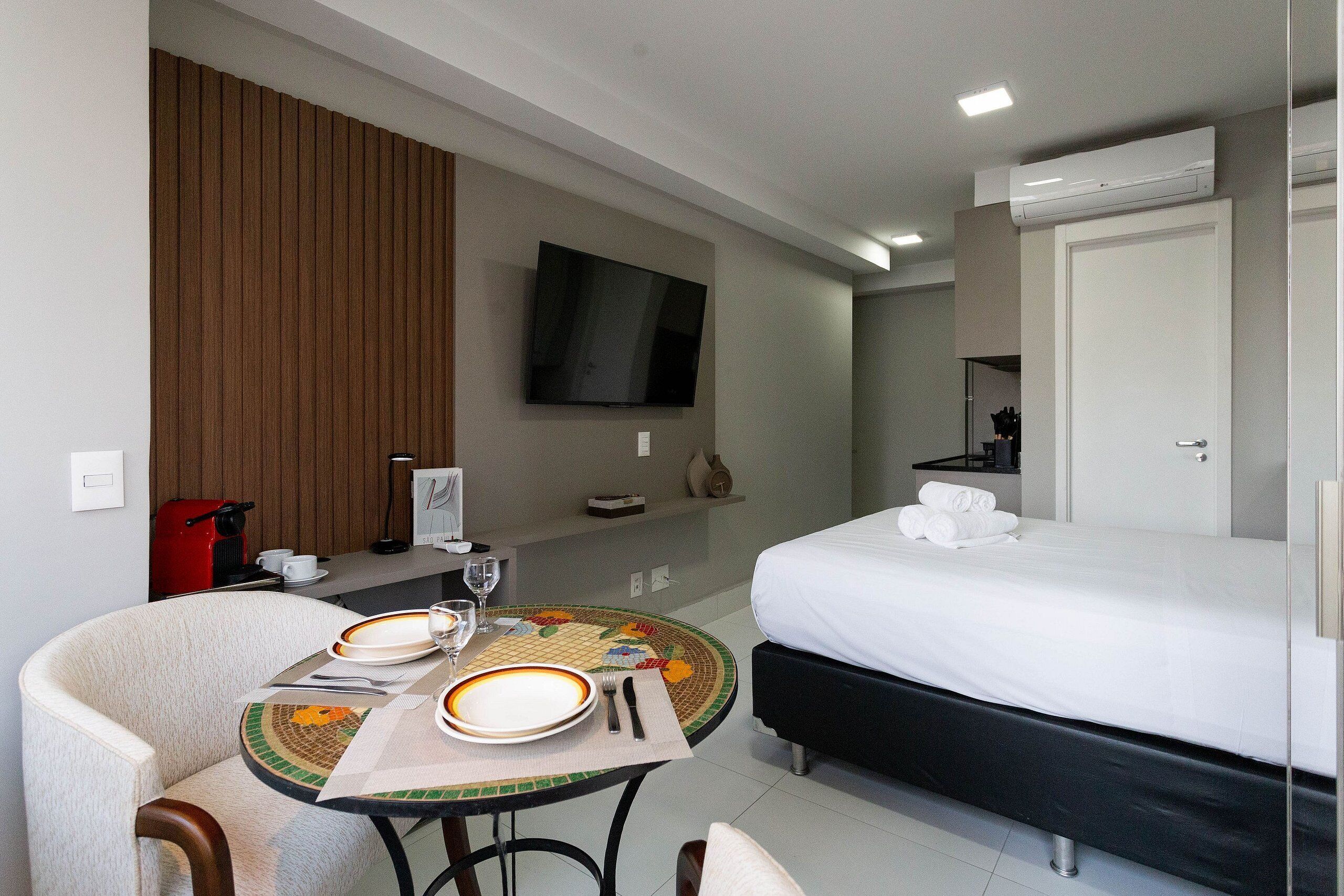 Una habitación de hotel con una cama, un televisor montado en la pared, una mesa de comedor con platos y cubiertos, y una silla.