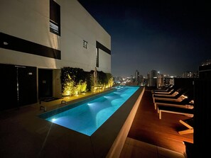 Una piscina en la azotea con vistas a la ciudad, rodeada de sillones de descanso y la fachada de un edificio moderno.