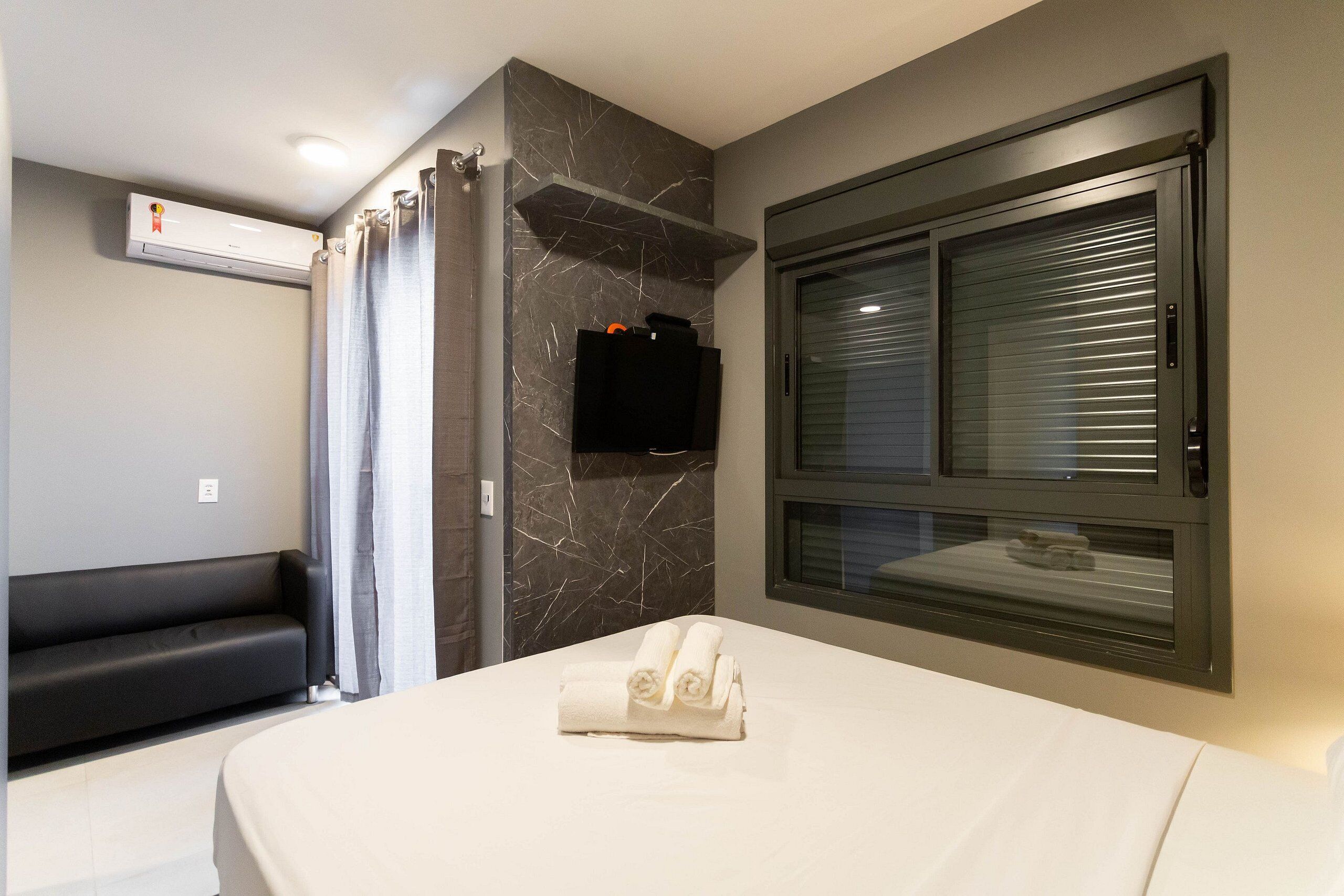 Una moderna camera d'albergo con un letto, una panca integrata, una TV a parete e una finestra con tende.