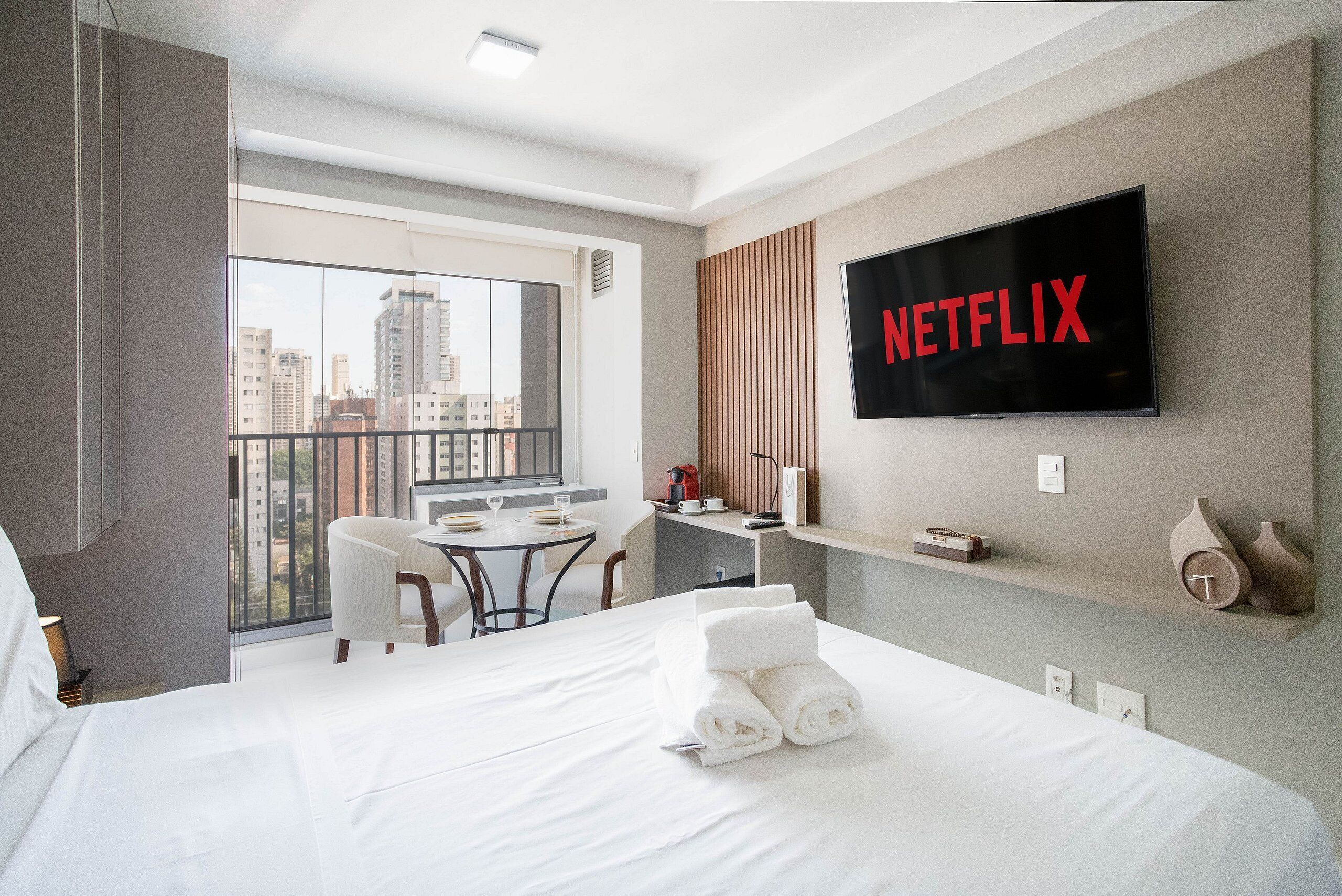 A hotel room with a large bed, a desk, a chair, a TV mounted on the wall, and a balcony with a city view.