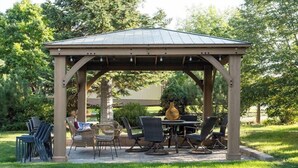 Un belvédère doté d’un toit soutenu par des poutres en bois, entouré d’une pelouse où se trouvent plusieurs chaises et une table.