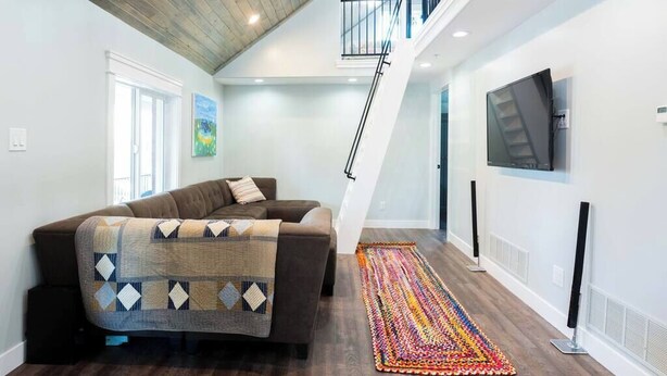 Un salon avec un canapé marron, un tapis coloré, un escalier et un téléviseur fixé au mur.