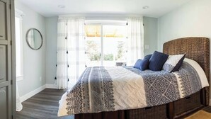 Une chambre avec un grand lit, une tête de lit en osier, une fenêtre avec des rideaux et un miroir sur le mur.
