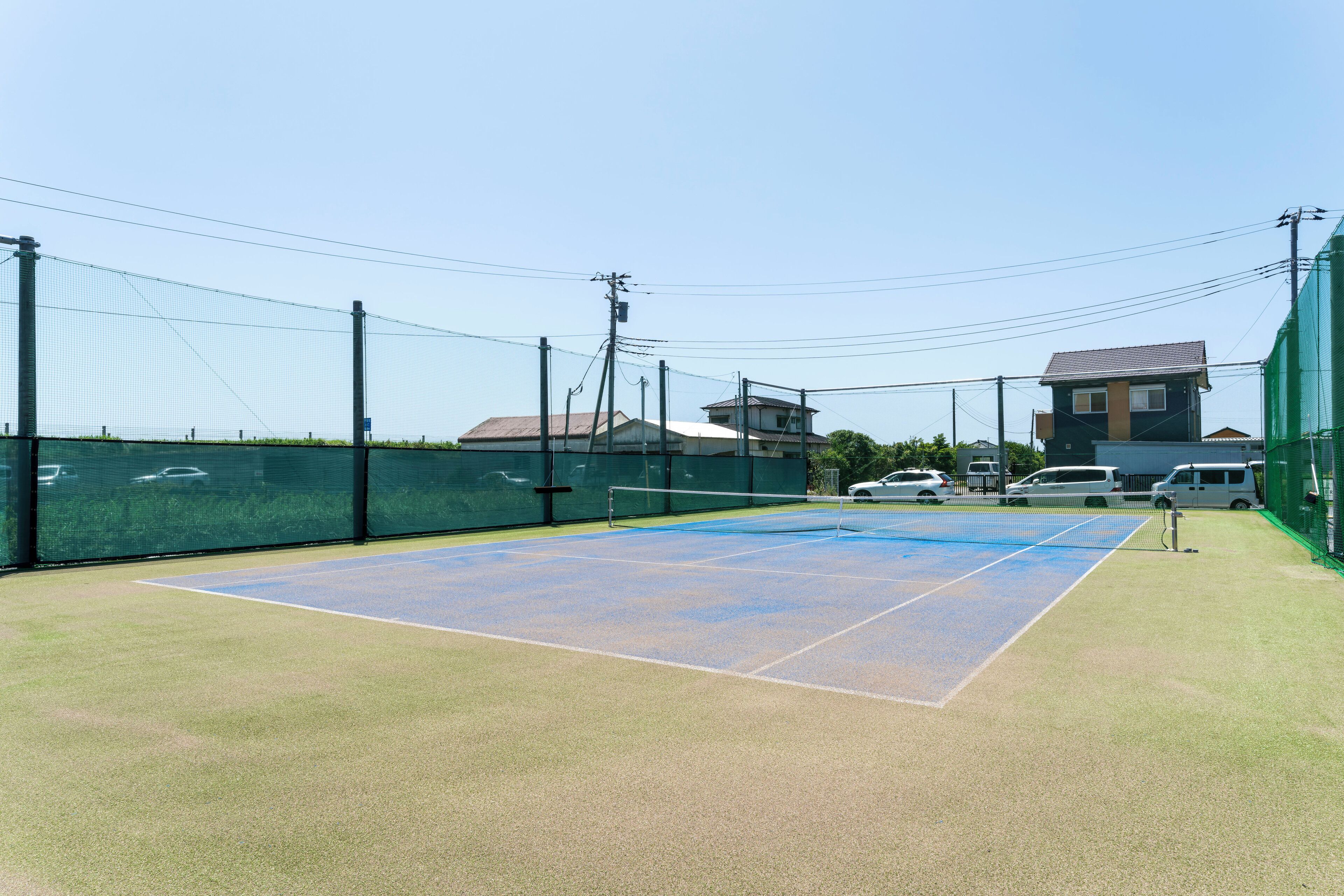 A tennis court with net, surrounded by a green fence, and buildings in the background.