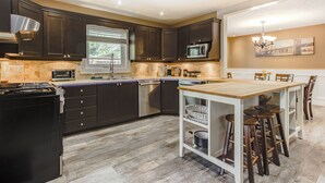 A modern kitchen with dark cabinets, stainless steel appliances, a large window, and a dining area with bar stools. - Amazing Lakeview Retreat with Hot Tub, Sauna and Private Loft! (Tiny)