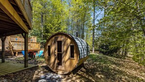 A wooden barrel-shaped structure with a door and windows, surrounded by a forested area with a wooden deck and outdoor seating. - Amazing Lakeview Retreat with Hot Tub, Sauna and Private Loft! (Tiny)