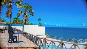 Une terrasse en bois avec des sièges extérieurs, offrant une vue sur une piscine et l’océan.