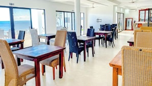 Une salle à manger spacieuse avec de hauts plafonds, de grandes fenêtres offrant une vue sur l’océan et des ventilateurs de plafond modernes.