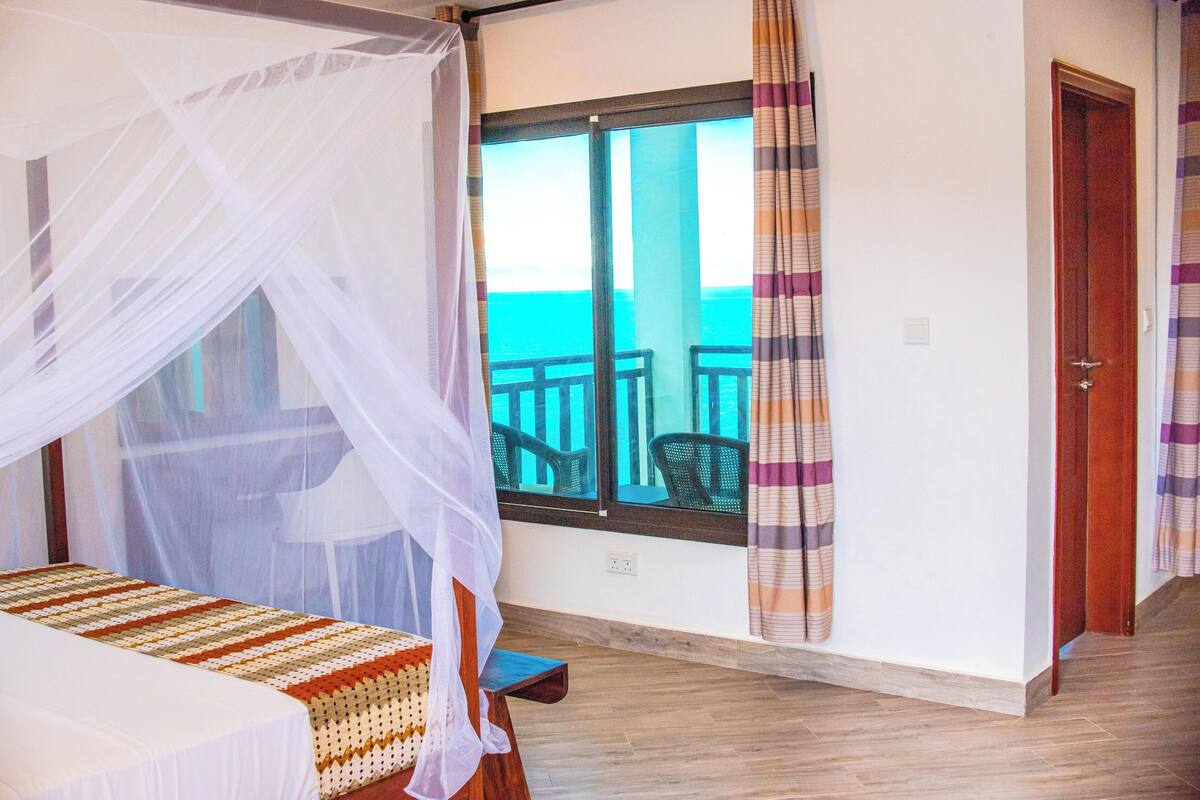 Une chambre avec un lit, une fenêtre donnant sur la mer, une porte et une petite table.