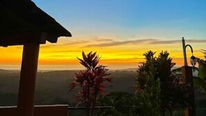 Vista do pôr do sol a partir de uma área externa coberta, com céu claro, palmeiras e uma cordilheira ao longe.