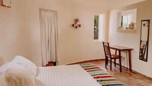 Quarto de hotel com cama, cadeira, mesa pequena e ventoinha no teto.