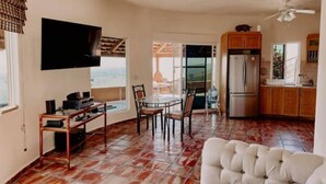 Uma sala de estar ampla com uma TV grande, uma mesa de jantar com cadeiras, um sofá branco e uma cozinha com armários de madeira e geladeira.