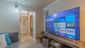Uma sala de estar com uma TV de tela plana exibindo diversos serviços de streaming, um sofá cinza e um rack de madeira para entretenimento.