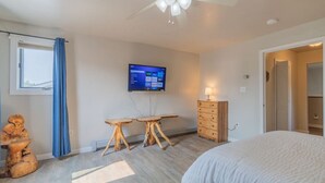 Quarto com cama, escrivaninha, penteadeira, ventoinha no teto e televisão na parede.