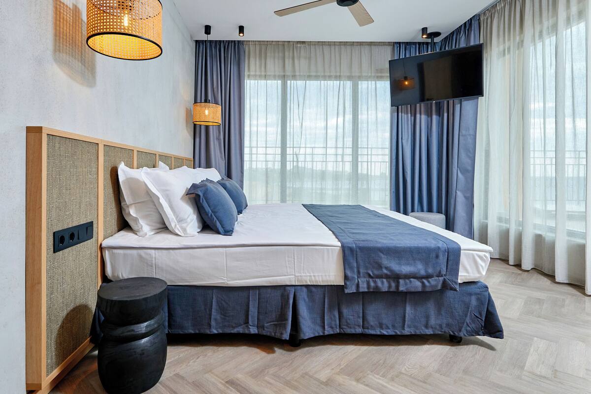A modern hotel room with a large bed, a flat-screen TV, and a view of the city through large windows.