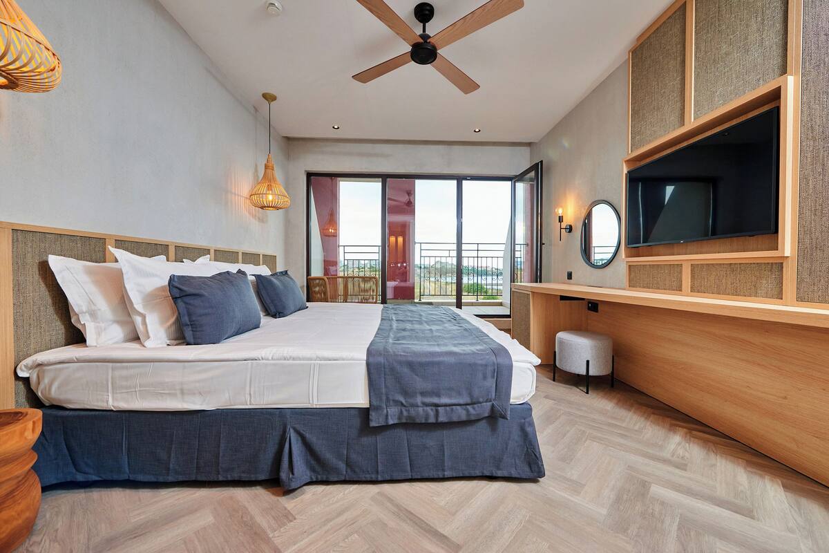A modern hotel room with a large bed, a ceiling fan, a TV mounted on the wall, and a balcony with a view.