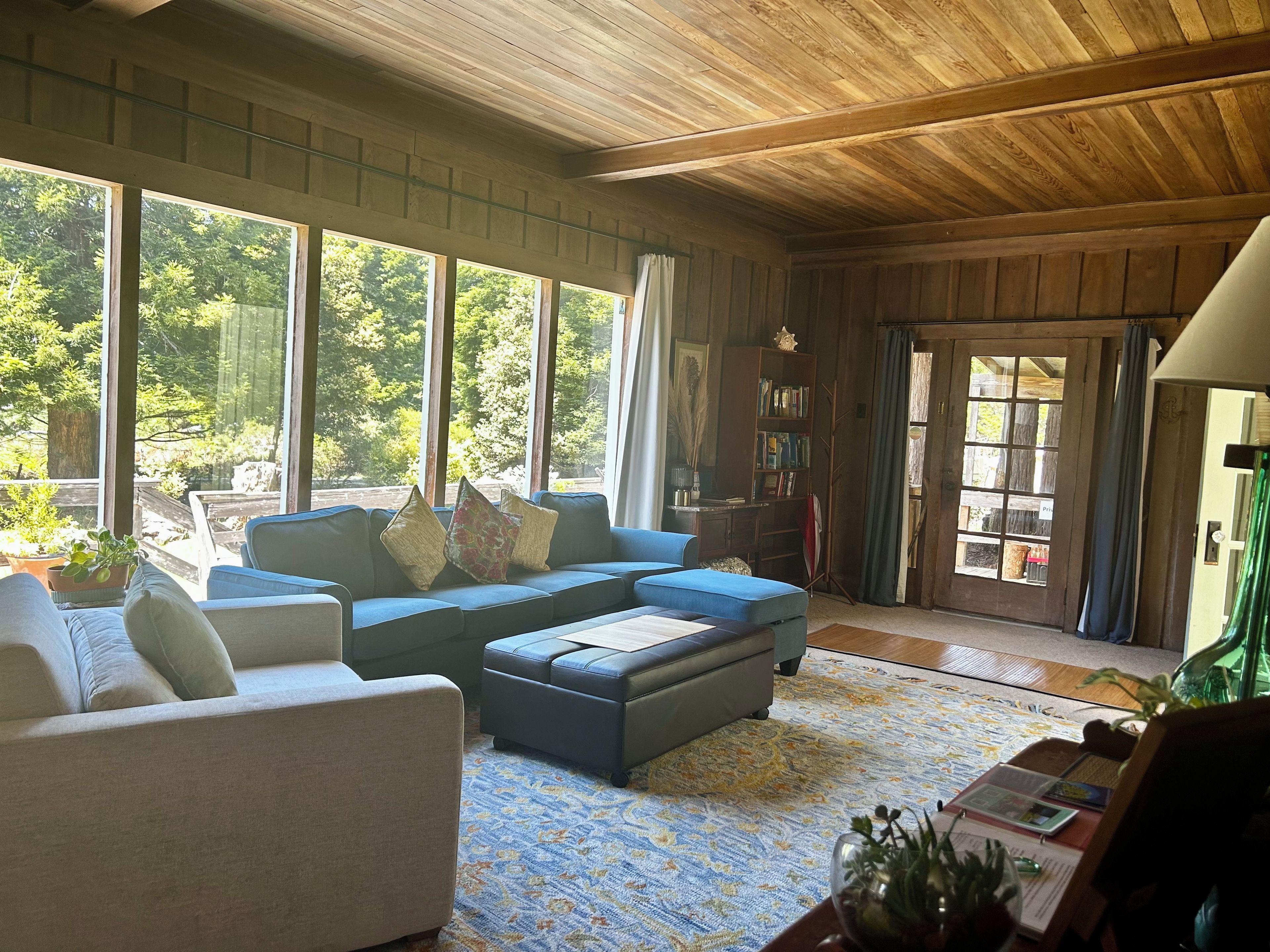 A spacious living room with a large window, a blue sofa, a white armchair, and a bookshelf.