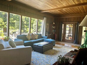 A spacious living room with a large window, a blue sofa, a white armchair, and a bookshelf. - Ranch home w/ hot tub in the Mendocino Redwoods. Pet- & family-friendly. (Fort Bragg)
