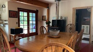Un coin repas avec une table en bois, des chaises, un saladier en verre et un poêle à bois.