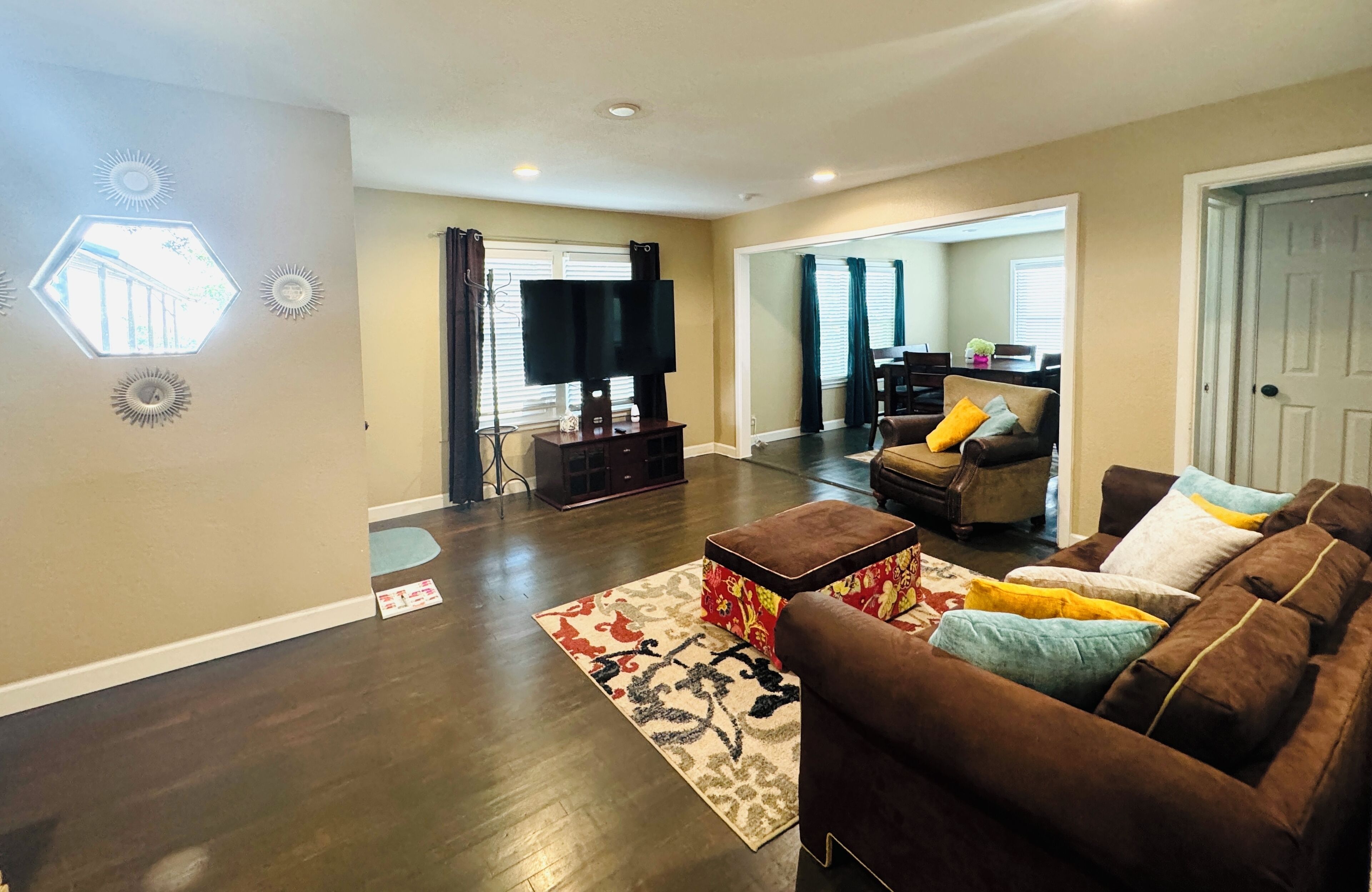 A living room with a brown sofa, ottoman, and a flat-screen TV. There is a dining area visible in the background.