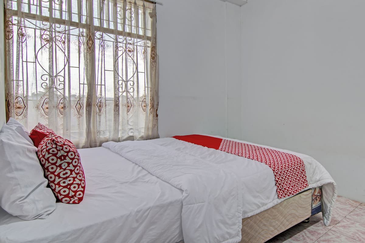 Un lit avec une literie blanche et une couverture à motifs rouges et blancs, un oreiller rouge et une fenêtre ornée de barreaux en métal décoratifs.