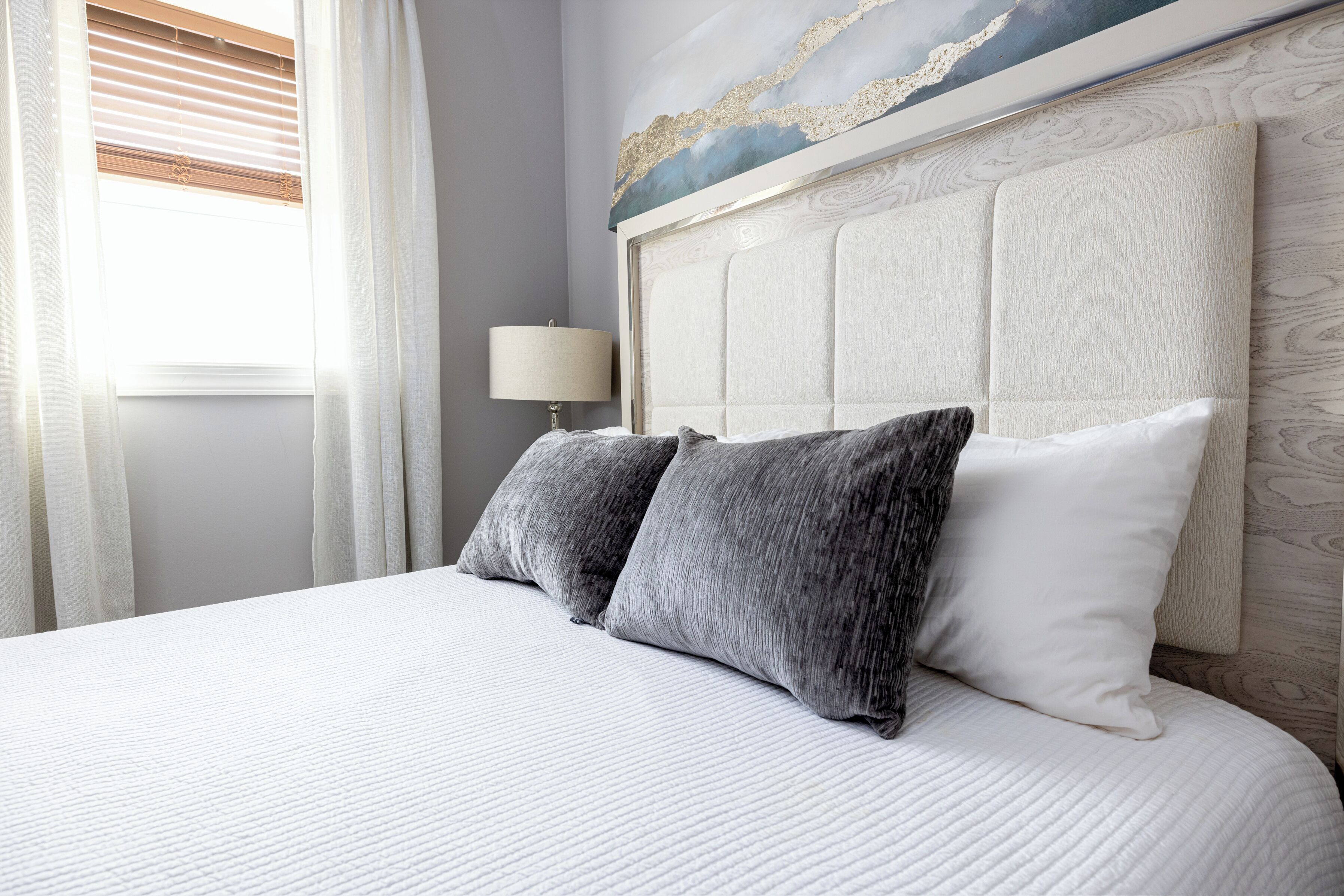 Un letto rifatto con coperte ordinate, un poggiatesta trapuntato, una lampada da comodino e un quadro sopra il poggiatesta.