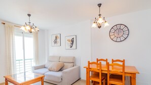 A living room with a sofa, a wooden dining table with chairs, a glass coffee table, and two wall-mounted light fixtures. There are two framed artworks on the wall and a round wall clock.