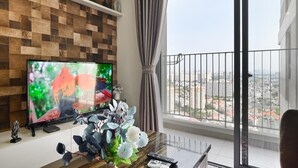 A modern living room with a large flat-screen TV, a wooden wall, a balcony with a view of the city, and a vase with flowers on the table. - Wonderful 2 bedrooms condo in T1 Masteri Thao Dien (Ho Chi Minh City)