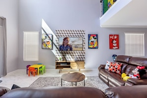 Una sala de estar con televisor, un sofá de cuero marrón, sillas coloridas, una mesa de centro de madera y un peluche de Minnie Mouse.