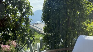 Un patio con una silla blanca acolchada, una mesa de mimbre con una planta en maceta, y una vista de una carretera y montañas al fondo.