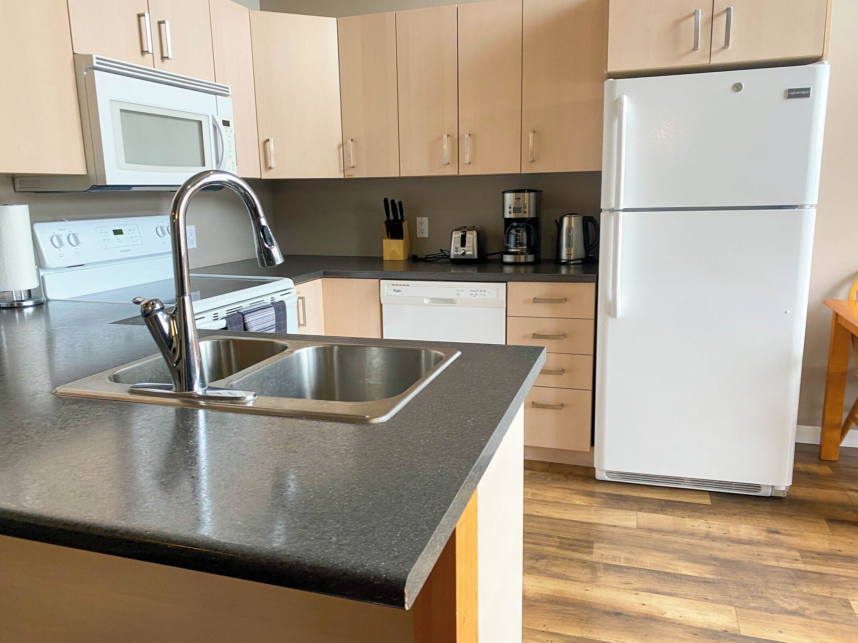 A modern kitchen with a stainless steel sink, a microwave, a refrigerator, and wooden cabinets.