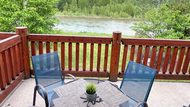 Eine Holzterrasse mit einem Glastisch und Stühlen, mit Blick auf einen Fluss und einen Wald.