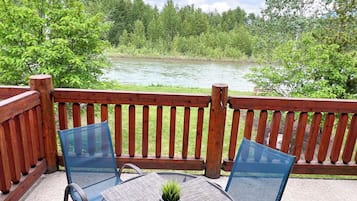 A wooden deck with a glass table and chairs, overlooking a river and forest.