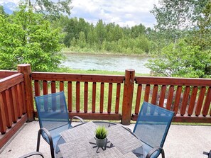 Eine Holzterrasse mit einem Glastisch und Stühlen, mit Blick auf einen Fluss und einen Wald.