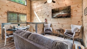 Un interno accogliente di una baita con una parete in legno, una ventola a soffitto, un tavolo da pranzo con sedie, un divano e un quadro appeso.