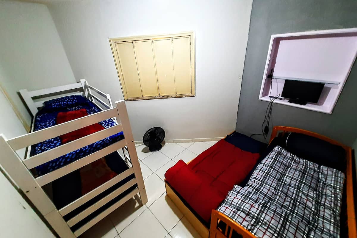 A dormitory room with bunk beds, a single bed, a window with blinds, and a small shelf with a television.