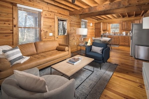 A cozy log cabin interior with a living room featuring a brown leather sofa, a blue armchair, and a coffee table. The kitchen area includes wooden cabinets and a stainless steel refrigerator.