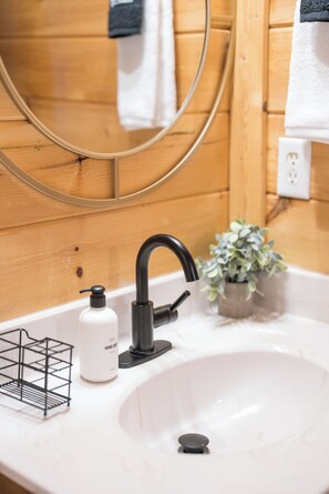 Ein Badezimmer mit einer Holzwand, einem weißen Waschbecken, einem schwarzen Wasserhahn, einem Spiegel, einer Topfpflanze und einer Handseifenspender.