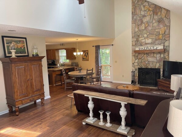 A spacious living room with a stone fireplace, a wooden dining table, and a kitchen in the background.